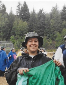 Shannon wearing a dark rain coat, dark green bucket hat, and a barely-there smile. She looks tired. There are pine trees in the background.
