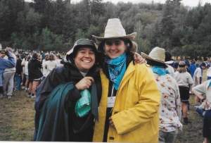 My sister and I smiling and standing side by side with arms around each other's backs. I'm in a black and blue rain jacket with a green bucket hat, my sister in a yellow rain jacket and straw cowboy hat. In the background are women milling about and scrubby pine trees.